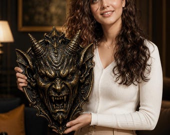May include: A decorative wall plaque featuring a detailed, three-dimensional sculpted demon face with prominent horns, pointed ears, and bared fangs. The piece is finished in a dark black color with metallic gold highlights that accentuate the texture.