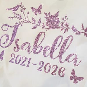 May include: A white fabric surface featuring a purple glitter design. The text reads Isabella 2021-2026 in a cursive font. Above the name is a decorative branch with a rose, leaves, and two birds. Three butterflies are positioned around the text.