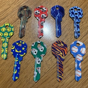 May include: A selection of patterned house keys. Designs include smiley faces, camouflage, footballs, lightning, hockey players, stars and moons, playing cards, tiger stripes, and baseballs. The keys are displayed on a wooden surface.