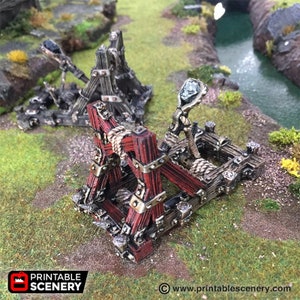 May include: Two miniature wooden trebuchets, one in the foreground with red and brown accents, the other in the background with grey and black tones. The trebuchets are set on a grassy surface, with a small stream in the background. The Printable Scenery logo is visible.