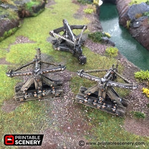 May include: Three wooden catapults with rope and metal accents. The catapults are painted in a weathered brown colour and have a rustic look. The catapults are set on a green grassy surface with a small stream in the background.