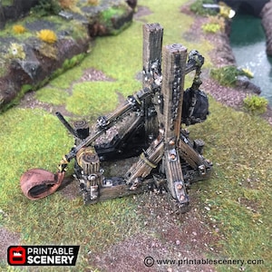 May include: A wooden catapult with a brown leather sling, a black metal arm, and a wooden base. The catapult is on a green grassy surface.