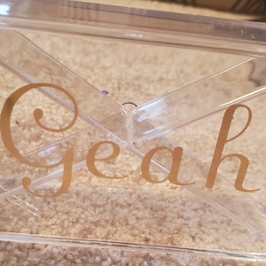 May include: A clear plastic box with a gold inscription that reads "Yeah".