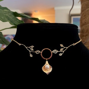 May include: A gold-colored necklace with a floral design. The necklace features a delicate chain with leaf accents and a central pendant. The pendant has a pearl and a gold-colored flower. The necklace is displayed on a black mannequin.