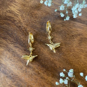 May include: A pair of gold hoop earrings with a hummingbird and star charm dangling from each hoop. The charms are adorned with small, clear rhinestones.