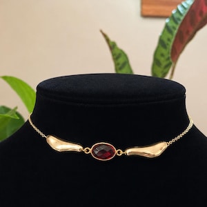 May include: A gold-coloured necklace featuring a central oval, deep red gemstone. The necklace has two asymmetrical gold accents either side of the gemstone, connected by a delicate chain. The necklace is displayed on a black mannequin.