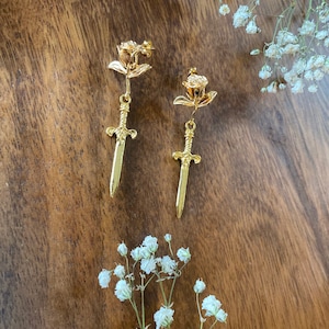 May include: A pair of gold-tone earrings featuring a delicate rose design with a sword dangling from the bottom.