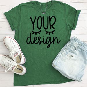 May include: A green t-shirt with the text "YOUR design" in black lettering. The design includes two stylized eyes. The t-shirt is laying on a white wooden surface with a pair of white sneakers and a pair of light blue denim shorts.