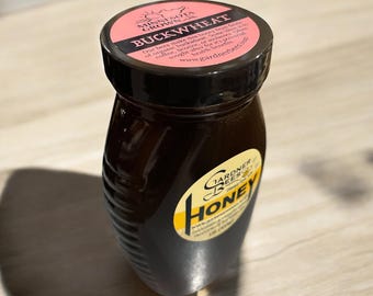 May include: A glass bottle of dark honey with a black lid and a pink label that reads "Minnesota Grown Buckwheat". The label also includes the text "Our bees make this honey from fields of organic buckwheat. Some say it tastes like coffee, licorice, or molasses. It's sought after for it's powerful health benefits. www.gardnerbees.com"