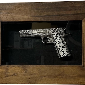 May include: A silver handgun with intricate floral engravings is displayed in a dark wood frame.