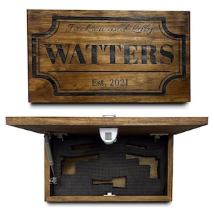 May include: A rectangular wooden wall-mounted storage box with a hinged lid. The front features the engraved text Jackson and Lilly, WATTERS, and Est. 2021. The interior contains dark grey foam inserts and a white latch mechanism.