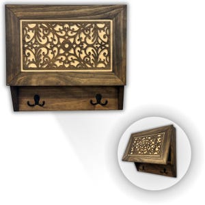May include: A wooden wall-mounted key holder with a decorative, openwork design. The dark brown frame surrounds a lighter-colored, intricately carved panel. Two black hooks are below. The key holder has a shelf and a hinged top.