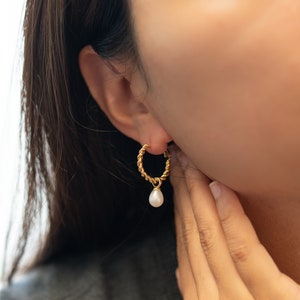 May include: Gold hoop earrings with a dangling pearl. The hoop is twisted and has a delicate design.