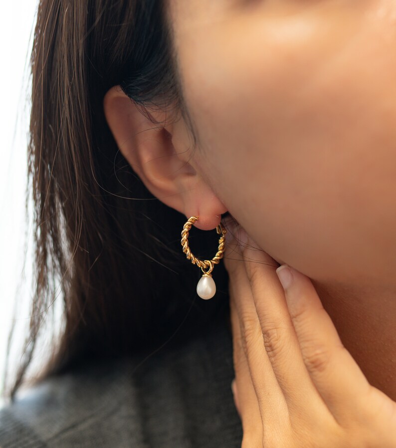 May include: Gold hoop earrings with a dangling pearl. The hoop is twisted and has a delicate design.