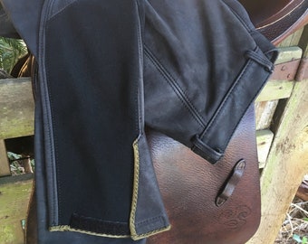 May include: A pair of black leather riding breeches with a dark green trim. The breeches are folded and placed on a brown leather saddle.
