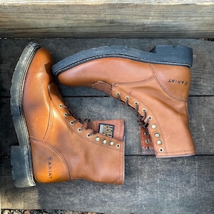May include: A pair of brown leather work boots with laces and a black sole. The boots have the Ariat brand name on the side.