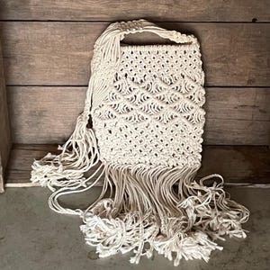 May include: A white crocheted bag with a long fringe. The bag has a woven pattern and a single strap.