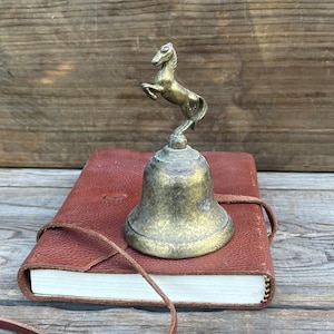 May include: A vintage brass bell with a rearing horse figurine on top, resting on a closed, red leather-bound journal. The bell and horse are aged with a patina finish. The journal has a leather strap closure.
