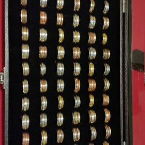 May include: A black velvet-lined box filled with a variety of silver, gold, and copper spinner rings. The rings are arranged in rows and columns, and each ring has a unique design.