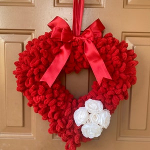 May include: A red heart-shaped wreath made of yarn with a red satin ribbon bow and white fabric flowers.