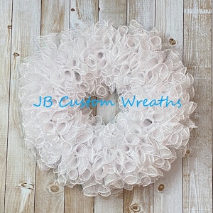 May include: A white wreath made of mesh with a ruffled texture. The wreath is on a wood background. The text "JB Custom Wreaths" is visible in the center of the wreath.