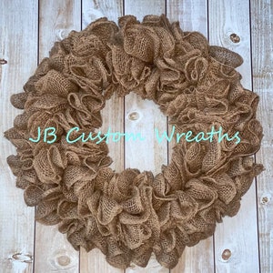 May include: A brown burlap wreath with a ruffled texture. The wreath is made of burlap mesh and is approximately 24 inches in diameter. The wreath is displayed against a white wood background.