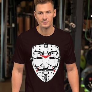 May include: A maroon t-shirt featuring a white graphic of a hockey mask with a stylised face and red accents. The mask resembles the one worn by the character "V" from the film "V for Vendetta".