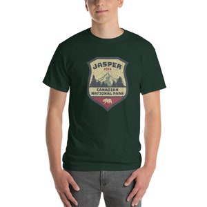 May include: A dark green t-shirt with a vintage-style logo for Jasper National Park in Canada. The logo features a mountain scene with a bear and the text "Jasper 2019 Canadian National Park".