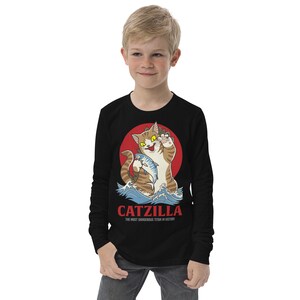 May include: Black long-sleeved shirt featuring a graphic of a cartoon cat holding a fish. The cat is standing in a wave with a red sun behind it. The text "CATZILLA THE MOST DANGEROUS TITAN IN HISTORY" is printed below the graphic.