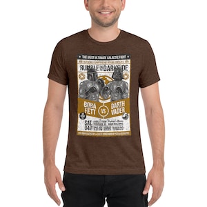 May include: A brown t-shirt with a vintage-style poster design featuring Boba Fett and Darth Vader boxing. The poster text reads "The Most Ultimate Galactic Fight" and "Rumble in the Darkside".