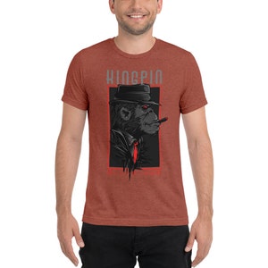 May include: A rust-colored t-shirt with a graphic of a black monkey wearing a fedora and a red tie, smoking a cigar. The text "KINGPIN" is above the monkey and the text "STAY TRUE" is below the monkey.