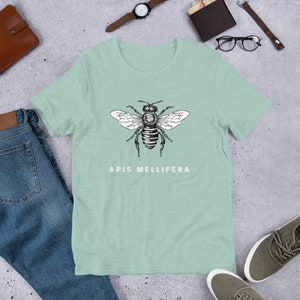 Puede incluir: Una camiseta verde claro con una ilustración en blanco y negro de una abeja y el texto "Apis mellifera" impreso en la parte delantera.