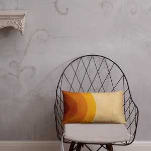 May include: A throw pillow with a retro-style geometric design in shades of orange, yellow, and brown. The pillow is sitting on a black wire chair with a white cushion.