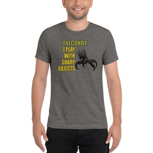 May include: A heather grey t-shirt with a black and white illustration of a bird's talon. The text "FALCONRY. I PLAY WITH SHARP OBJECTS" is printed in yellow on the shirt.