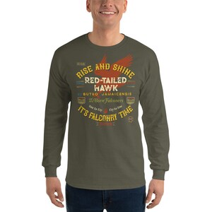 May include: A green long-sleeved t-shirt with a vintage-style graphic design featuring a red-tailed hawk, the text "Rise and Shine", "Red-Tailed Hawk", "Buteo Jamaicensis", "We are Born Falconers", "Live to Fly", "Fly to Live", and "It's Falconry Time".