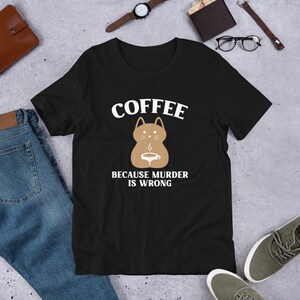 Puede incluir: Camiseta negra con un gráfico blanco de un gato marrón sosteniendo una taza de café. El texto "COFFEE BECAUSE MURDER IS WRONG" está impreso debajo del gato.