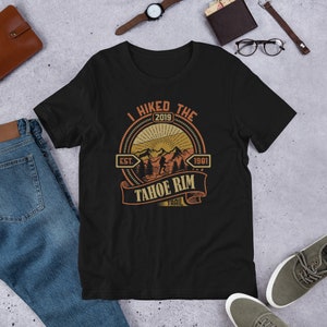 Op de afbeelding: Zwart T-shirt met een vintage-stijl grafische afbeelding met de tekst "I Hiked The Tahoe Rim Trail Est. 1981 2019". De afbeelding toont een bergketen met een wandelaar en een zon.