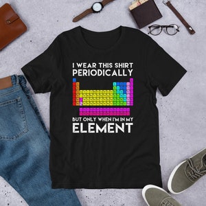 Op de afbeelding: Zwart T-shirt met een kleurrijke afbeelding van het periodiek systeem en de tekst "Ik draag dit shirt periodiek, maar alleen als ik in mijn element ben."