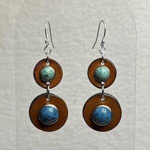 May include: A pair of handcrafted earrings featuring brown leather circles with turquoise and blue stone accents. The earrings have silver-colored hooks and a layered design, showcasing a bohemian style.
