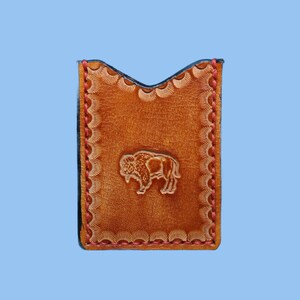 May include: A tan leather card holder featuring a stamped bison design in the center. The edges are finished with a decorative scalloped border and secured with red stitching. The top edge has a V-shaped cutout for easy card access.