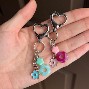 May include: Two silver-colored heart-shaped clasp keychains with flower charms. Each keychain has a chain with three colorful flower charms in shades of blue, pink, and purple. The keychains are held in a hand.