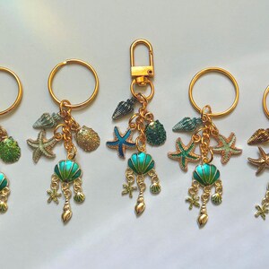 May include: Gold keychains with ocean-themed charms. Each keychain features a gold ring or clasp, adorned with starfish, seashells, and a teal and gold pendant. The charms are connected by gold chains.