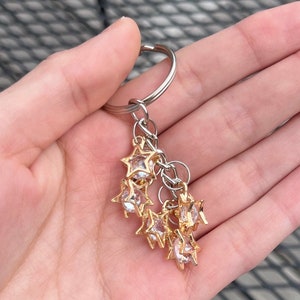May include: A keychain featuring a silver ring and a cluster of gold-coloured star charms. Each star charm has a clear, faceted centre. The keychain is held in a hand.