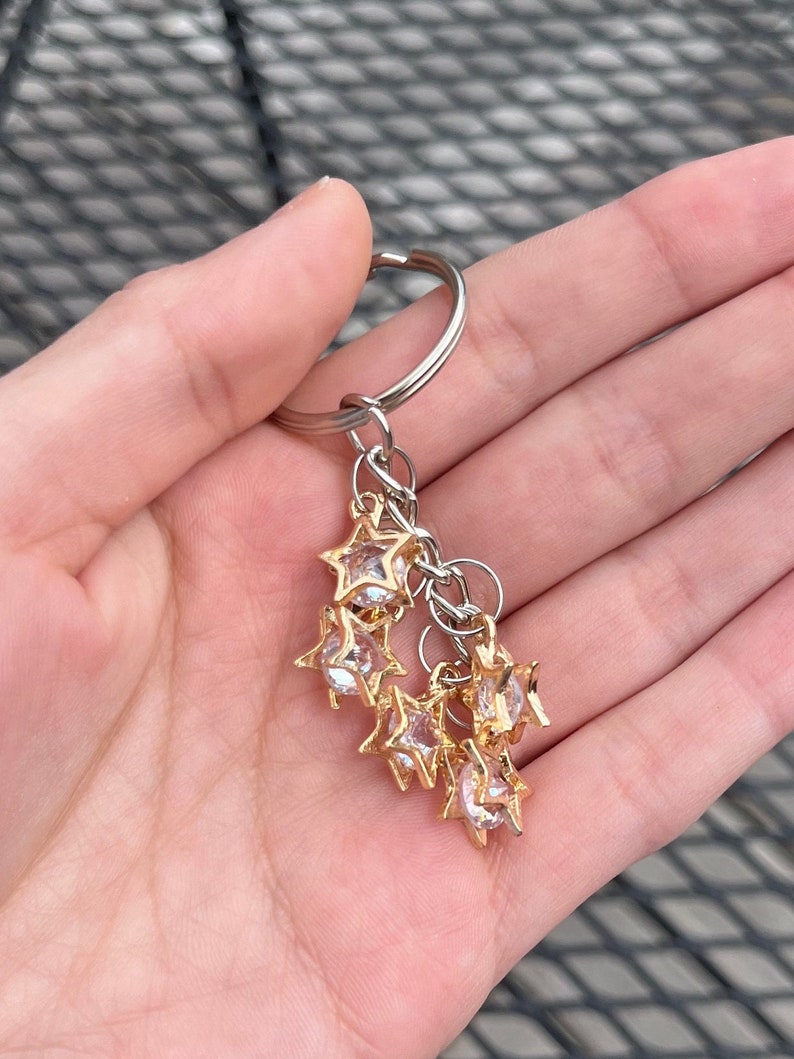 May include: A keychain featuring a silver ring and a cluster of gold-coloured star charms. Each star charm has a clear, faceted centre. The keychain is held in a hand.