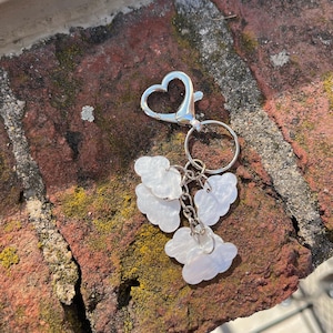 Puede incluir: Un llavero plateado con un cierre en forma de corazón y un anillo que sostiene varios dijes en forma de nube. Los dijes son blancos y nacarados, unidos con pequeñas cadenas. El fondo es una pared de ladrillo desgastada.