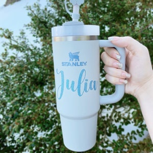 May include: A white Stanley travel mug with a gray handle and a blue glitter name "Julia" on the side. The mug has a white flower-shaped lid with a straw.
