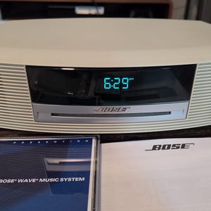 May include: A beige Bose Wave music system with a digital clock display showing 6:29 AM, a CD slot, and the Bose logo. Included are a CD case labeled Presenting The Bose Wave Music System and a white instruction manual with the Bose logo.