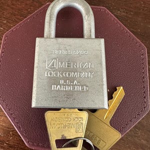 May include: A silver-colored metal padlock with a hardened shackle, featuring the embossed text SERIES 5200, AMERICAN LOCK COMPANY, U.S.A., and HARDENED. Two brass-colored keys are attached to the lock, each marked with the American Lock Company logo.
