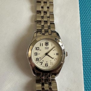 May include: A silver-toned Swiss Army wristwatch featuring a round white dial with black Arabic numerals, a date window at the three o'clock position, and a metallic link bracelet. The watch face displays the Swiss Army logo and text.
