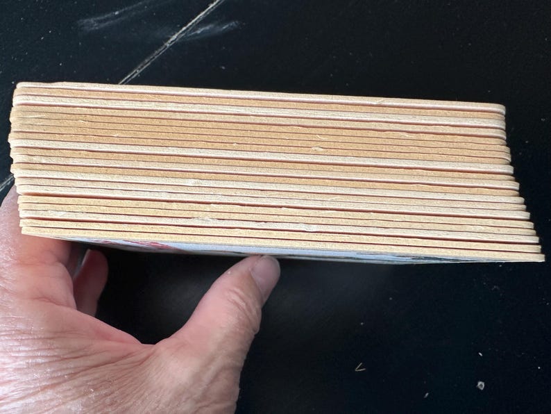 Puede incluir: Una pila de posavasos rectangulares de madera con esquinas redondeadas. Los posavasos son de color marr&oacute;n claro con una fina l&iacute;nea marr&oacute;n rojizo cerca de los bordes. La pila est&aacute; sostenida por una mano.